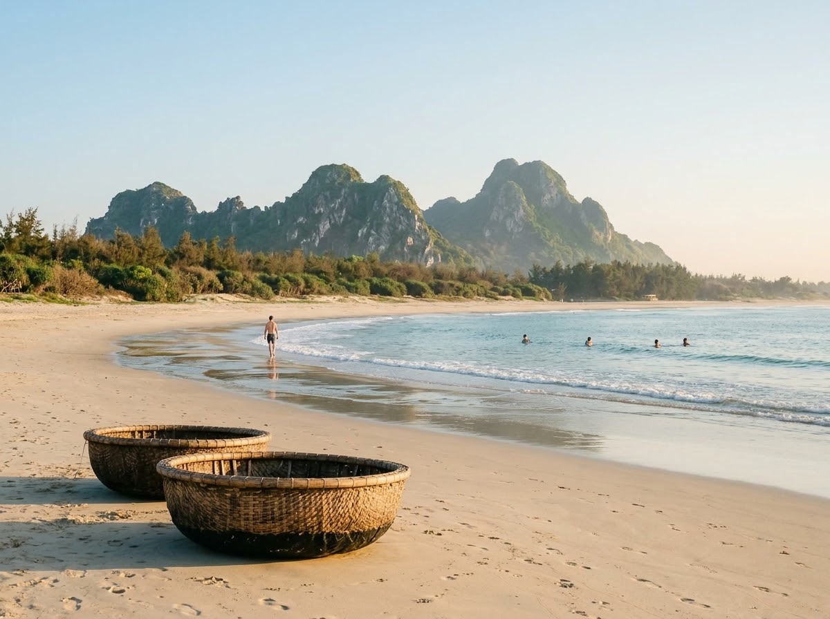 Early-morning view along a wide pale-sand beach in Da Nang under a clear sky, gentle low waves, a few swimmers in the water and a beachgoer walking at the water line, round woven basket boats resting on the sand, and the rounded green-grey limestone outcrops of the Marble Mountains rising behind low trees