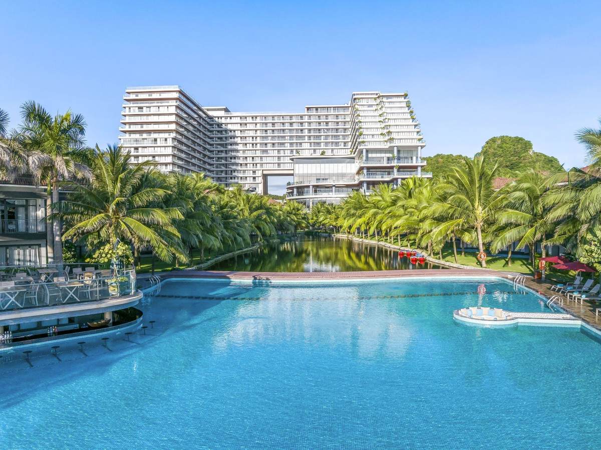 Large blue pool with a swim-up bar and stools on the left, a palm-lined waterway behind and a tall white building beyond