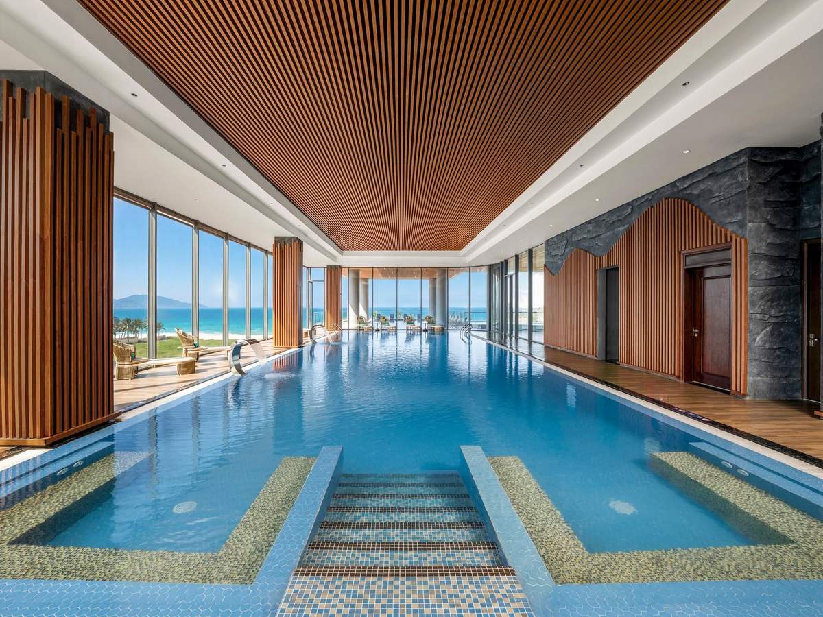 Long indoor pool under a slatted wooden ceiling, with floor-to-ceiling windows looking over the sea