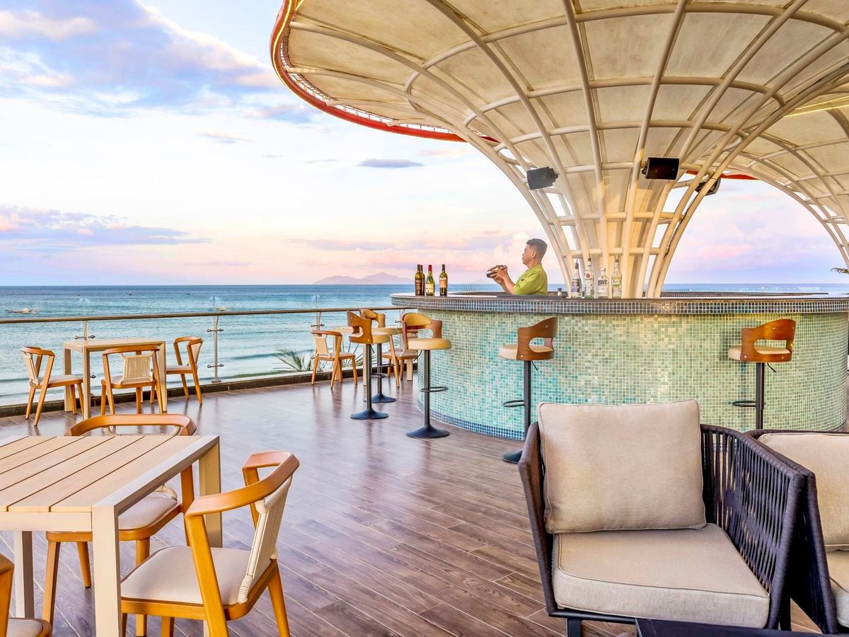 Open-air bar deck with a round mosaic counter under a white canopy, tables and the sea at sunset