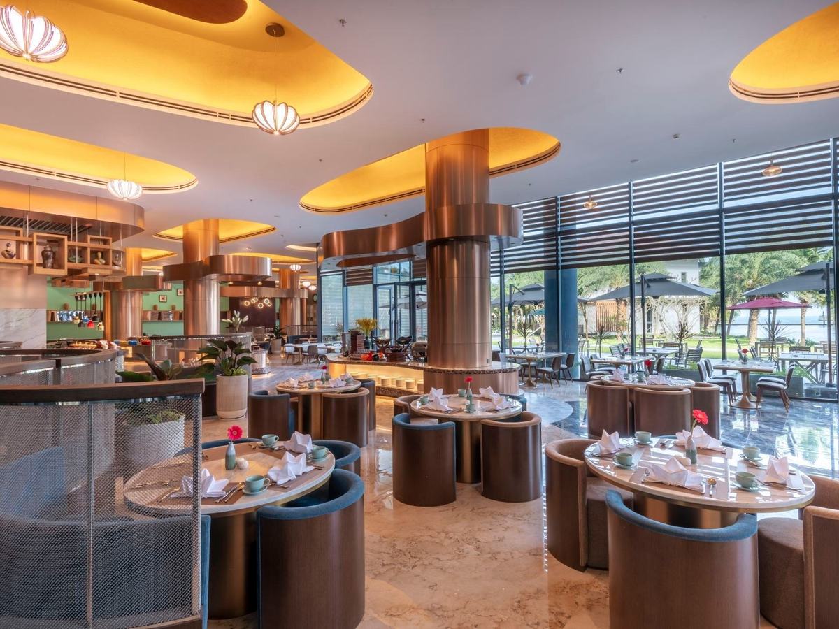 Restaurant with round marble tables, bronze columns and pendant lamps, and glass walls opening onto a garden terrace