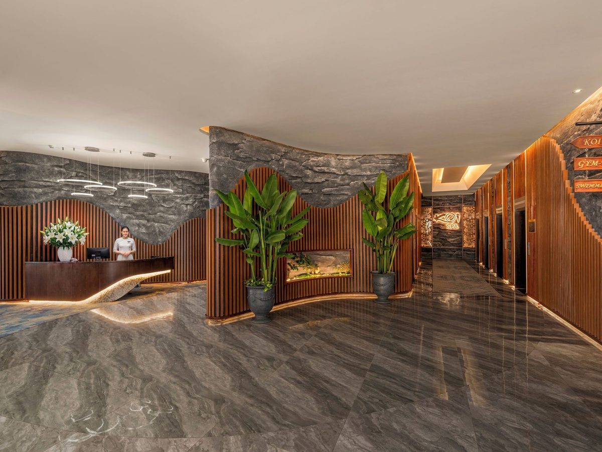 Spa reception with a curved wooden desk, stone-textured walls, tall potted plants and direction signs on the wall