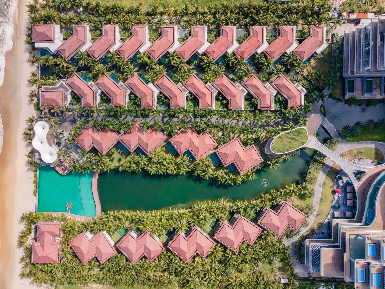 Aerial view of rows of red-roofed villas around a green lagoon and a turquoise pool beside a sandy beach